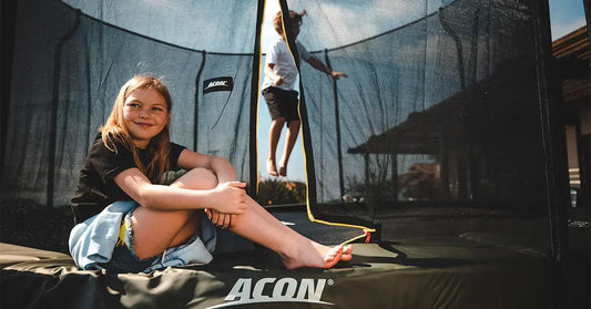 Jente som sitter på kanten av en Acon-trampoline mens en gutt hopper inni den.